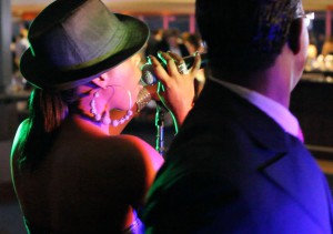 A woman wearing a hat and large hoop earrings sings into a microphone, while a man in a suit—part of the Boston wedding band—stands in the foreground facing away.