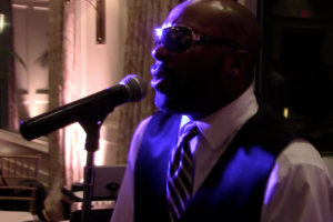 A man wearing sunglasses, a dress shirt, tie, and vest sings into a microphone under stage lights, bringing the energy of a top Boston wedding band to the stage.
