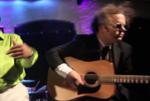 A man in a suit and sunglasses from a Boston wedding band plays an acoustic guitar on stage next to another person in a bright green shirt, with colorful stage lighting in the background.