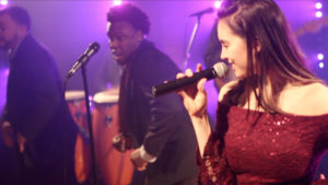 A woman sings into a microphone on stage with a Boston wedding band; two men stand near her and drums are visible in the background under stage lights.