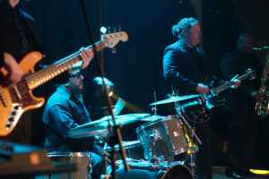 A Boston wedding band performs on stage with musicians playing the bass guitar, drums, electric guitar, and saxophone under vibrant stage lighting.