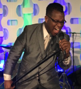 A man in a gray suit passionately sings into a microphone on stage, leaning forward and gripping the mic stand, as music equipment hums in the background—capturing the energy of a top Boston wedding band performance.