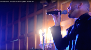 A man in a dark suit holds a microphone and sings indoors, with warm lights and wood-paneled walls in the background—capturing the lively spirit of a Boston wedding band performance.
