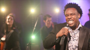 A male singer in a suit passionately performs into a microphone on stage as his Boston wedding band plays instruments behind him, illuminated by bright stage lights.
