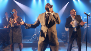 A Boston wedding band performs on stage with blue lighting; a man in a suit sings into a microphone, flanked by a keyboardist, a woman backup singer, and a man playing tambourine.