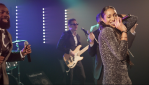 A Boston wedding band performs on stage with a female lead singer in a sparkly dress, male vocalist, bassist, and drummer, all illuminated by colorful stage lights.